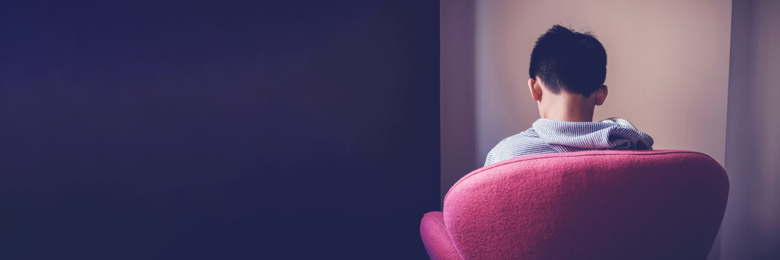 A sad autistic boy is sitting in a dark room with his back facing us.